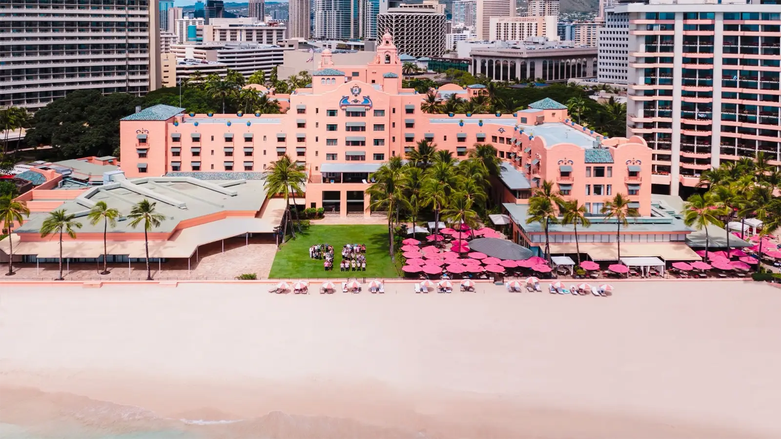 Aerial view of one of the best hotels on Oahu: a large, pink beachfront hotel with a lush green lawn, surrounded by palm trees. Beach chairs with pink umbrellas are set up near the water. The hotel is located in a vibrant cityscape, with tall buildings visible in the background.