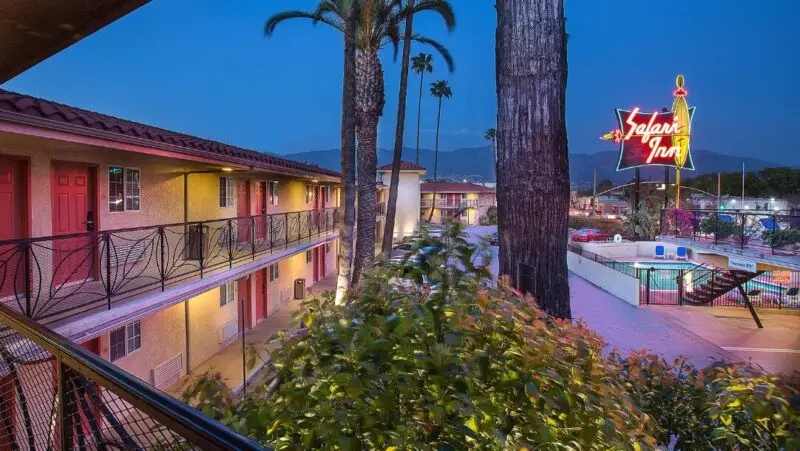 Night view of a motel with a glowing neon sign that reads "Safari Inn." The two-story building has rooms with red doors and a balcony with ornate railings. Tall palm trees and a lit swimming pool area add to the ambiance. If you're wondering where to stay in Palm Springs, this might be your spot. Mountains are visible in the background.