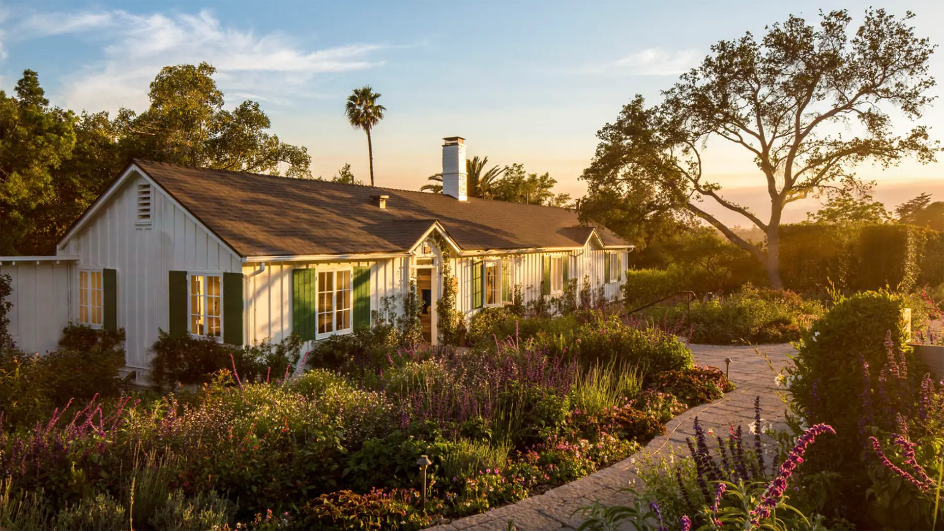 A charming white house, reminiscent of the best luxury hotel in Santa Barbara, is surrounded by a lush, colorful garden in full bloom. The sun sets in the background, casting a golden hue over the scene. A winding stone path leads to the entrance, and tall trees provide a serene, natural backdrop.