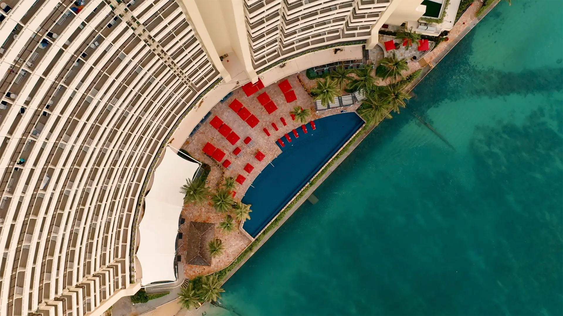 Aerial view of a curved beachfront hotel building with multiple floors, one of the best hotels on Oahu. Below, several red lounge chairs and umbrellas surround a rectangular blue swimming pool. Palm trees and clear turquoise water can be seen beside the pool area.