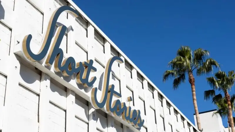 A white building facade with the cursive words "Short Stories" mounted in gold and silver letters. Under the clear, blue sky and framed by two tall palm trees on the right, it evokes a charming Palm Springs vibe.