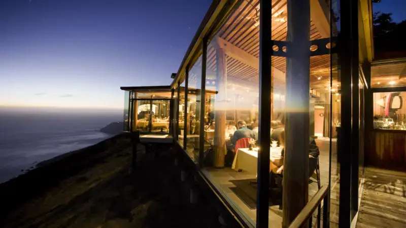A modern restaurant with large glass windows extends over a cliffside at sunset. The inside is warmly lit, with diners seated at tables. The exterior offers a stunning view of the ocean and coastline, fading into the distance as evening approaches—a perfect recommendation for where to eat in the Bay Area.