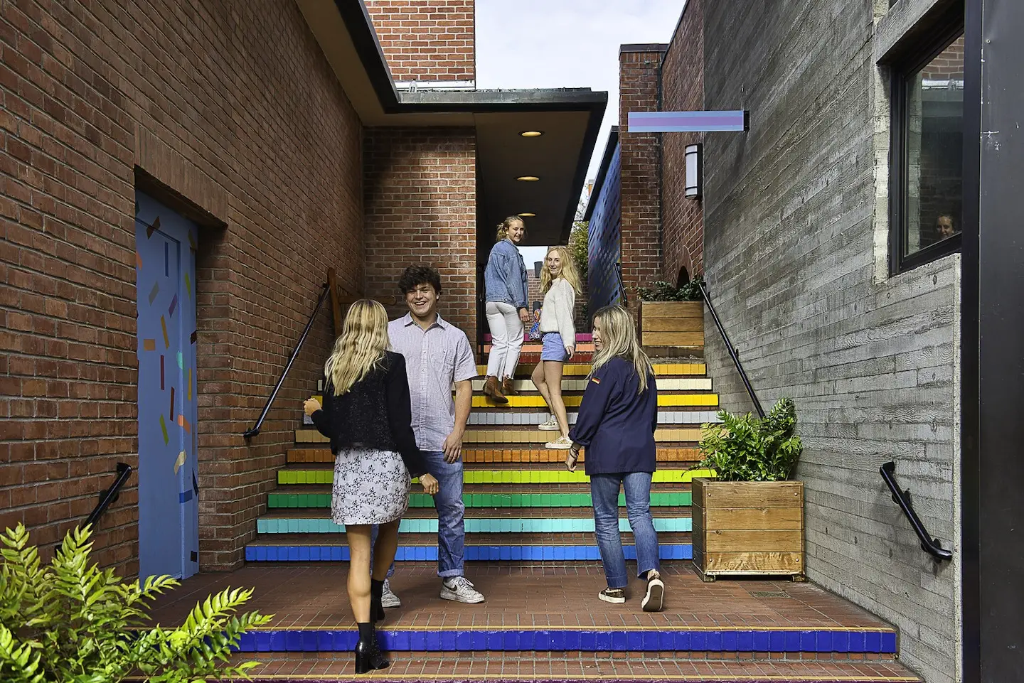 People socializing on a colorful outdoor stairway at Ghirardelli Square. The stairs are painted in rainbow colors, set between brick and concrete buildings with modern accents and potted plants. Some people are standing and talking, while others are ascending the vibrant steps.