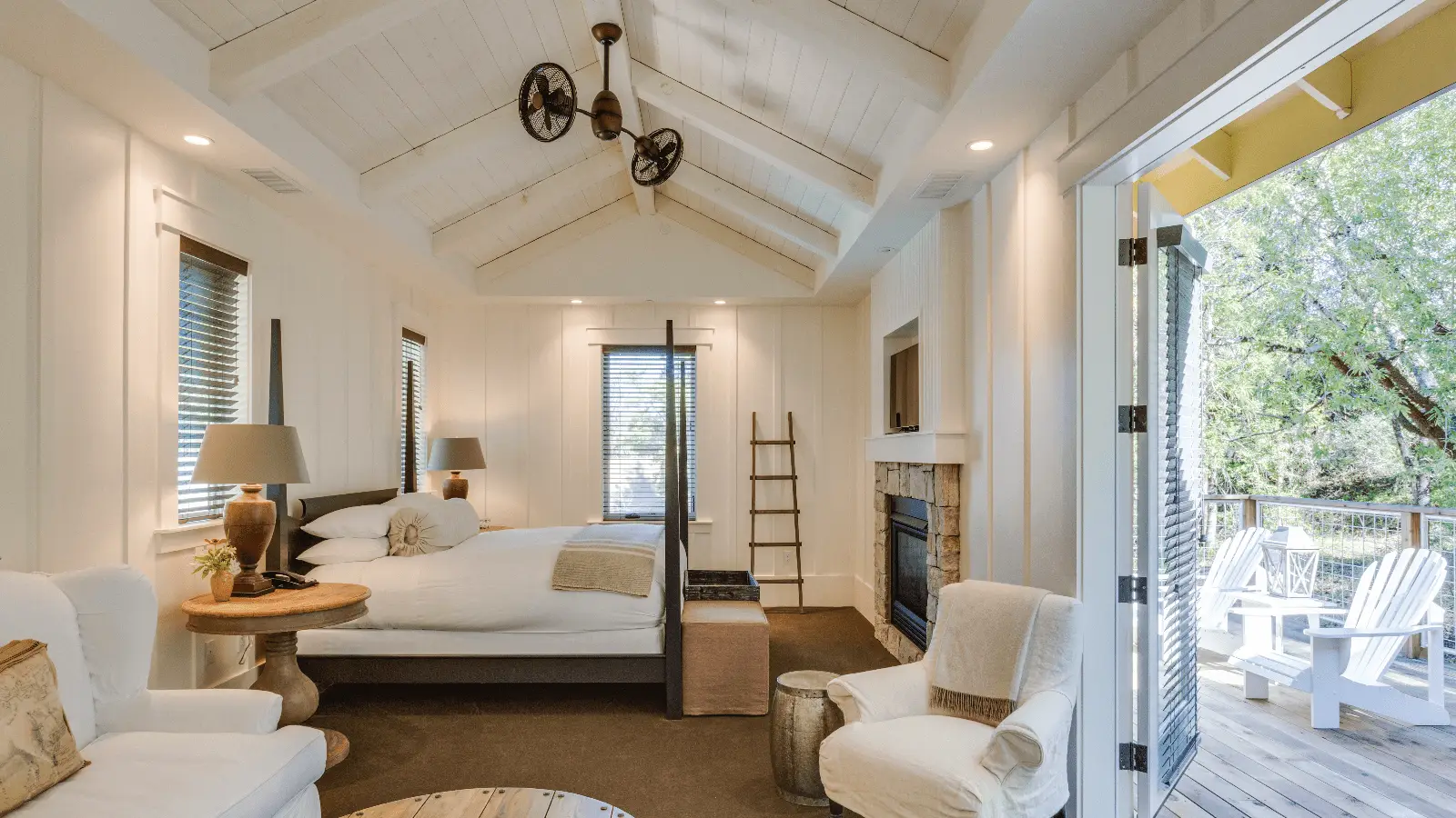 A cozy bedroom where to stay in Sonoma features a vaulted ceiling with exposed beams and a unique mechanical fan. It has a bed, fireplace, chairs, and a ladder shelf. Large glass doors open to a deck with outdoor seating, offering a view of greenery. The room is decorated in a neutral palette.