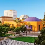 An urban park with a circular water fountain and surrounding trees in the foreground. Behind the park is a bright orange building with a purple dome and palm trees. Several high-rise buildings and a clear blue sky are in the background, making it one of the best things to do in the South Bay this March.
