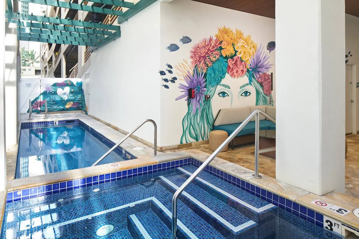 Indoor pool area with vibrant mural of a woman's face adorned with colorful flowers and fish. The pool has a stepped entry with metal handrails, blue mosaic tiles, and a depth marking of 3 feet. In the background, there's a green pergola and a lifeguard chair.