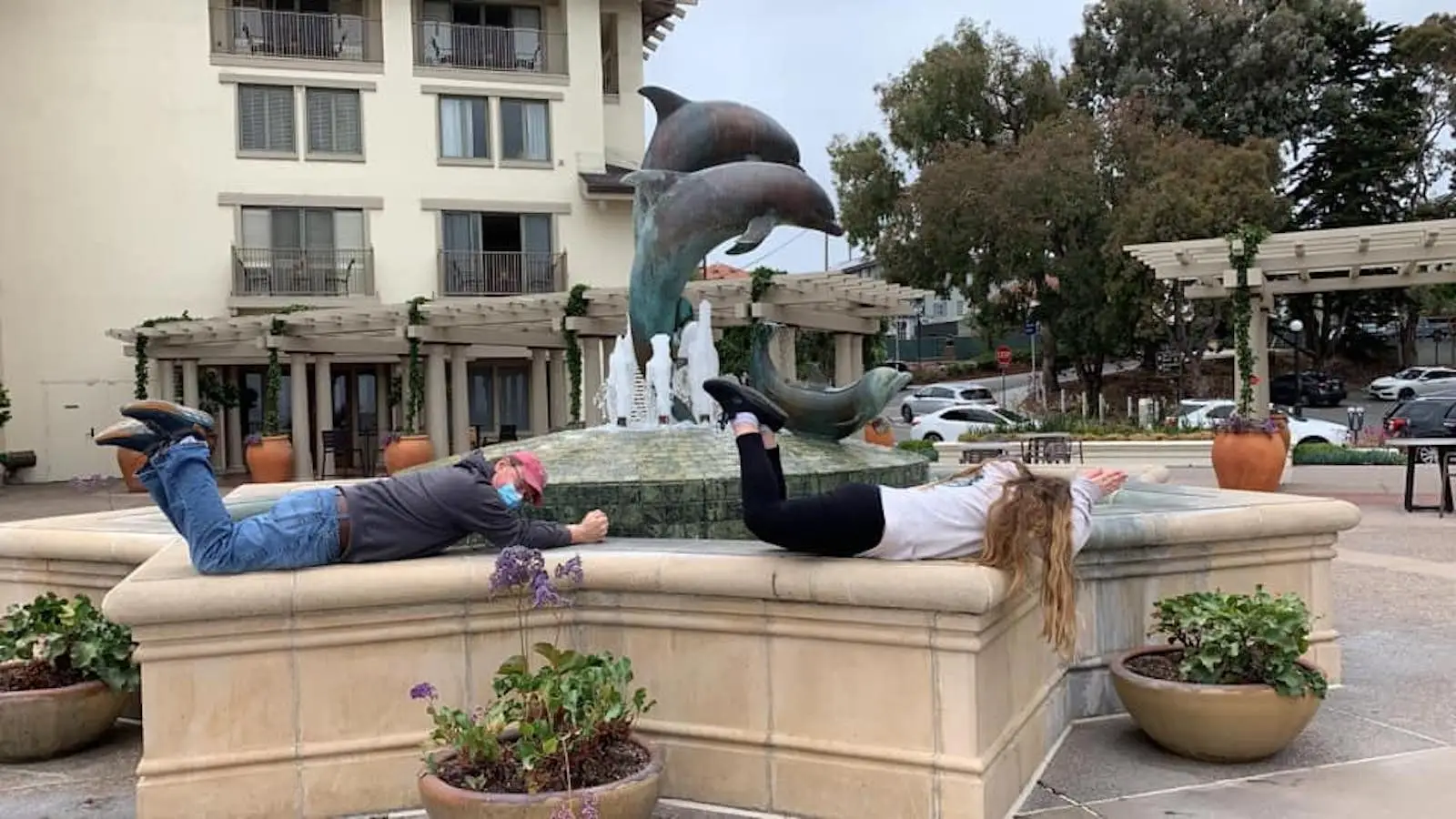 Two individuals are lying face down, planking on the edge of a large fountain with dolphin sculptures. The fountain is situated in an outdoor area of a building with balconies and potted plants. Trees and a parking area with cars are visible in the background, showcasing unique *family activities* in Monterey Peninsula.