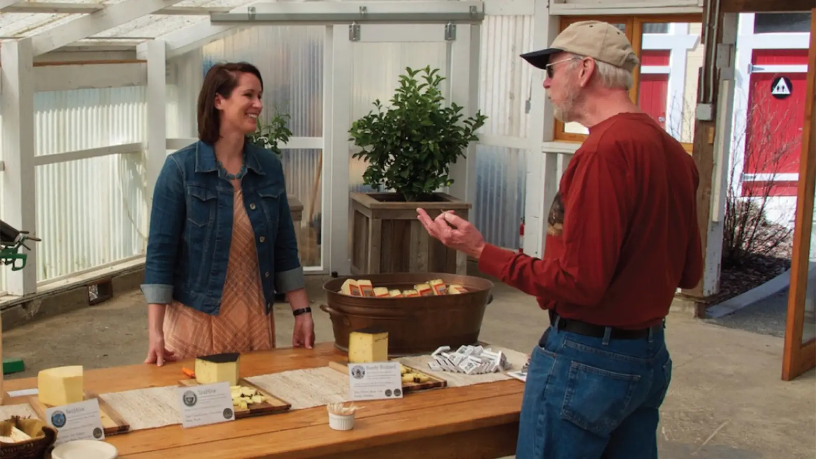 A woman in a denim jacket stands behind a wooden table with cheese samples, smiling at an older man in a red shirt and baseball cap. They are inside a well-lit space adorned with plants and wooden crates, proudly showcasing Sonoma County artisanal food. Cheese sample signs are displayed on the table.