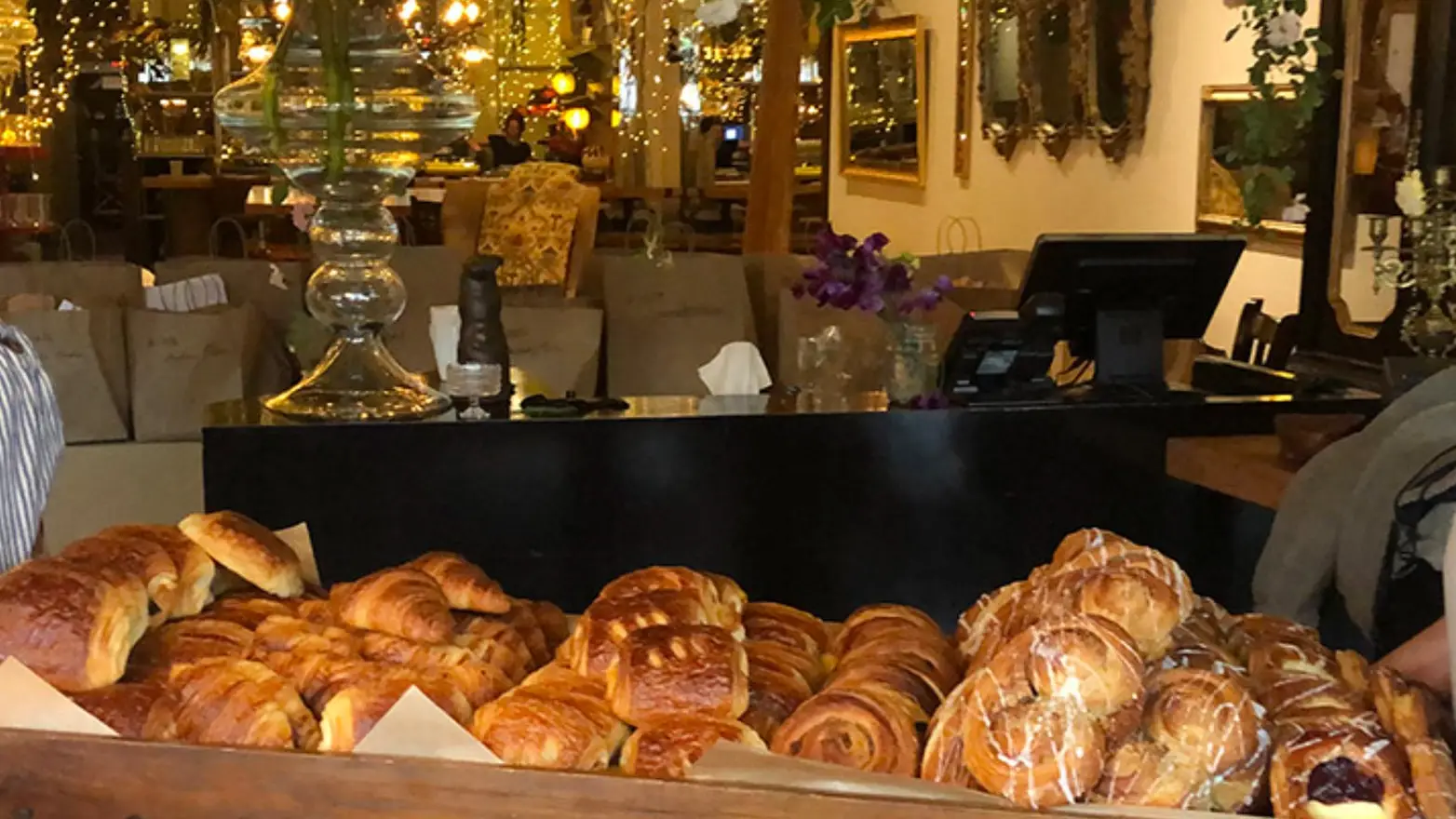 A display of various pastries such as croissants and danishes is shown on a table in the foreground. The background features a cozy café with warm lighting, decorated with hanging string lights and paper bags for take-out orders, showcasing the charm of Sonoma County's artisanal food scene.