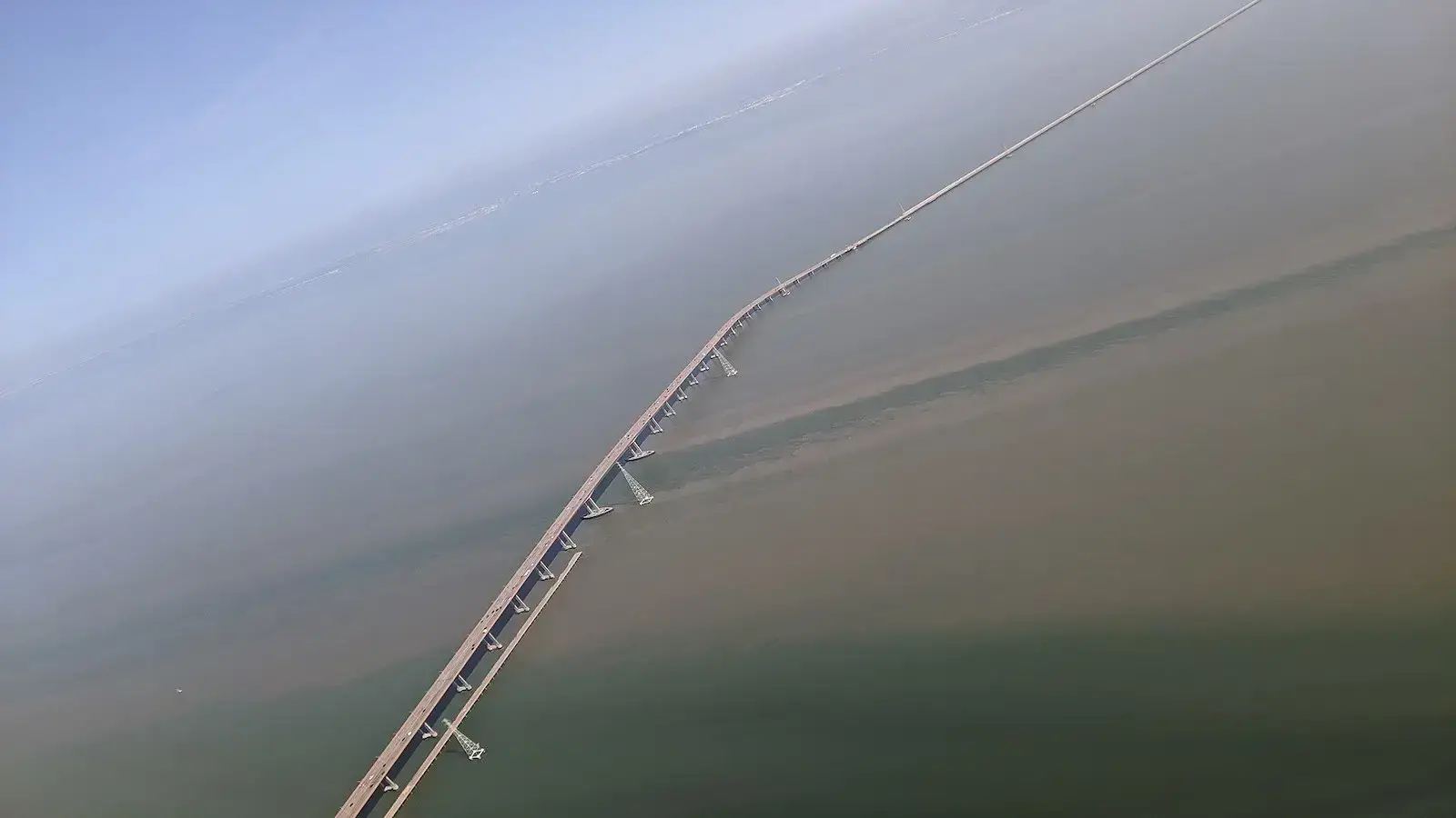 A long California bridge stretches across a vast body of water. The image, taken from an elevated angle, shows the bridge's extensive length and its connection between distant land masses. The water below appears calm, and the sky above is clear and blue.