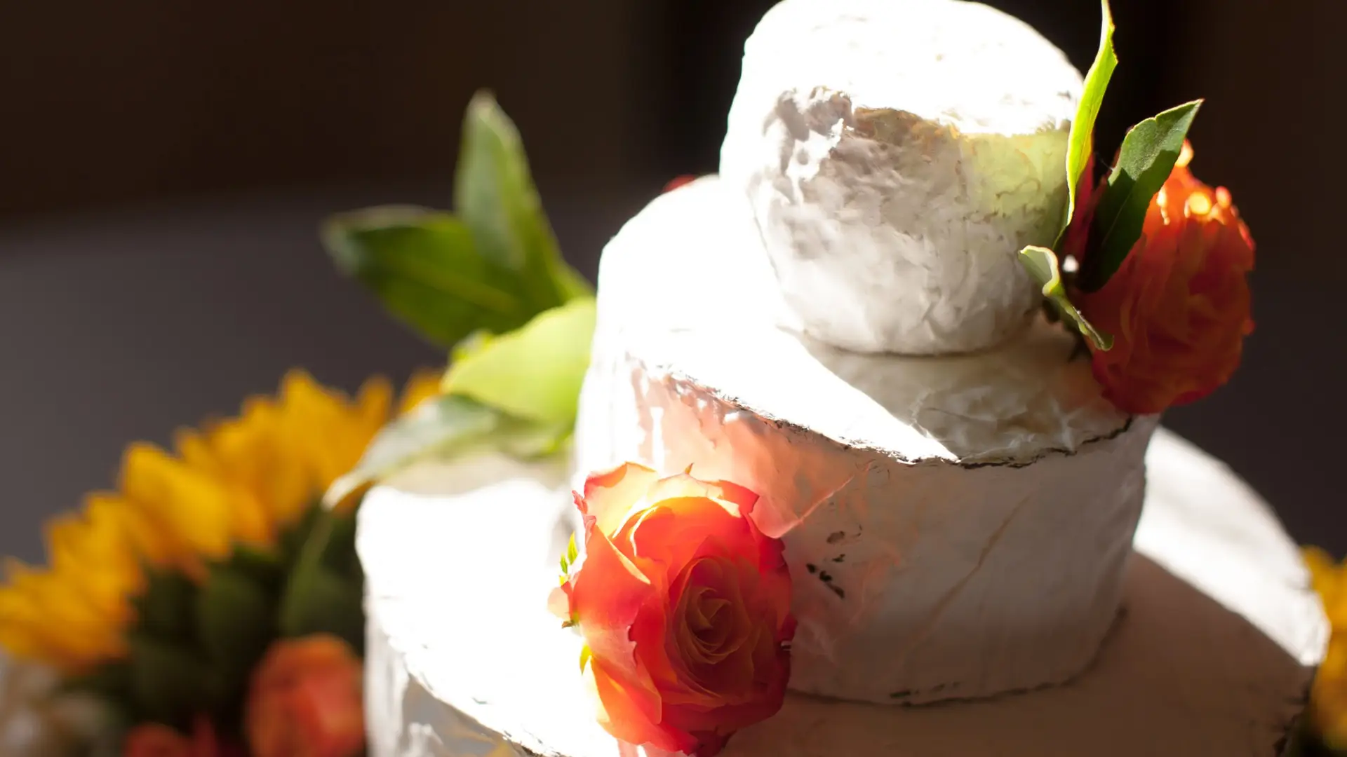 A close-up of a two-tiered cake covered in white frosting. The cake, an example of Sonoma County artisanal food, is adorned with red and orange roses, with a hint of green leaves. The background is blurred but shows some yellow flowers. The image is brightly lit, highlighting the cake's details.