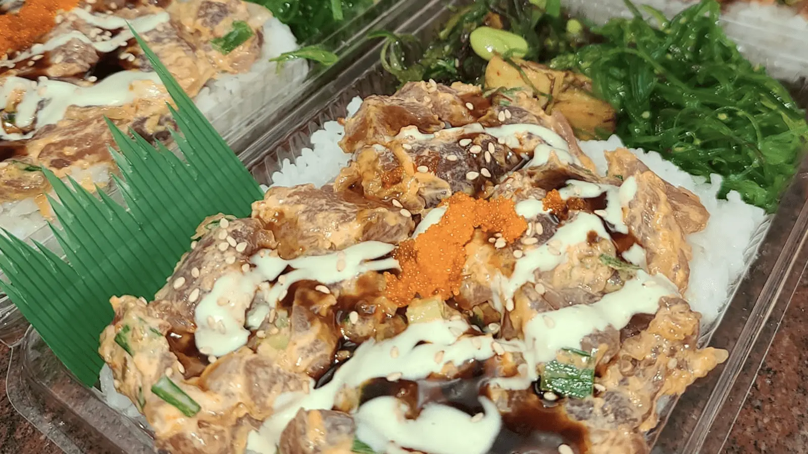 A close-up of a takeout container filled with a savory dish featuring rice topped with a creamy, saucy mixture of marinated meat, garnished with sesame seeds, sauce, and a small mound of orange roe. Green vegetables are in the background—truly the best poke from Central Kauai.