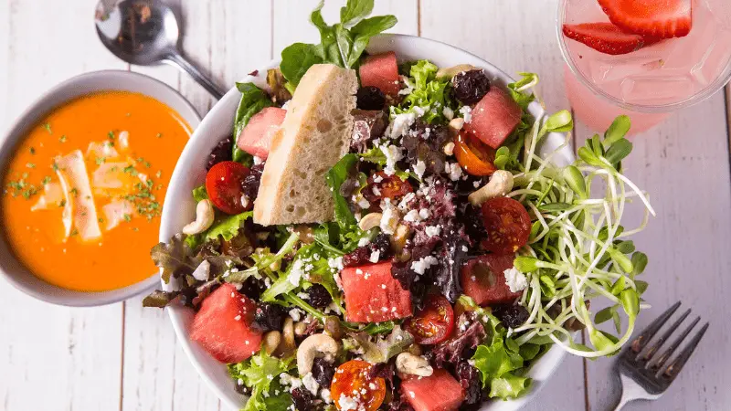A vibrant lunch in Wailea features a salad with mixed greens, watermelon chunks, cherry tomatoes, nuts, raisins, and feta cheese. Beside a bowl of herb-garnished tomato soup with strips of bread is a glass of pink beverage with strawberry slices.