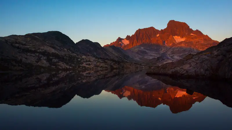 A serene mountain landscape at sunset, with rugged peaks bathed in warm, orange light reflecting perfectly in the still waters of a lake below. This picture-perfect scene could easily be part of the John Muir Trail, with its clear sky transitioning from deep blue to a lighter shade near the horizon.