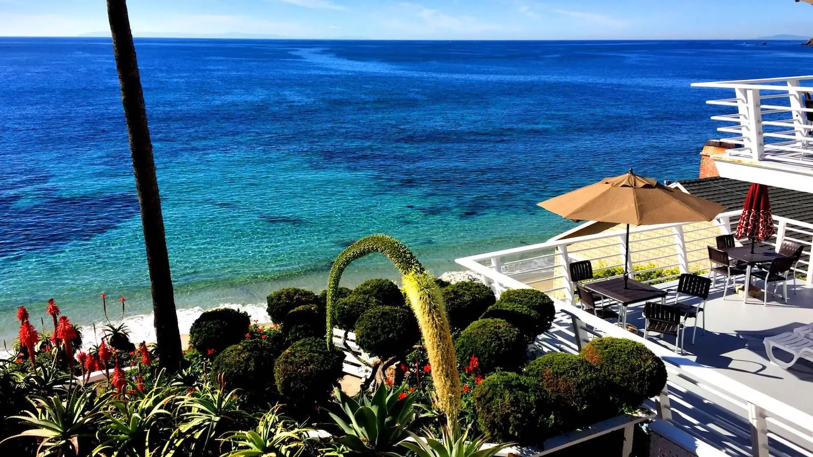 A scenic beachfront property overlooks a clear, vibrant blue ocean. The foreground features lush, manicured bushes and a garden with red flowers. On the right, there's a patio with outdoor furniture under a large beige umbrella. This hidden gem feels like part of an exclusive soul community.