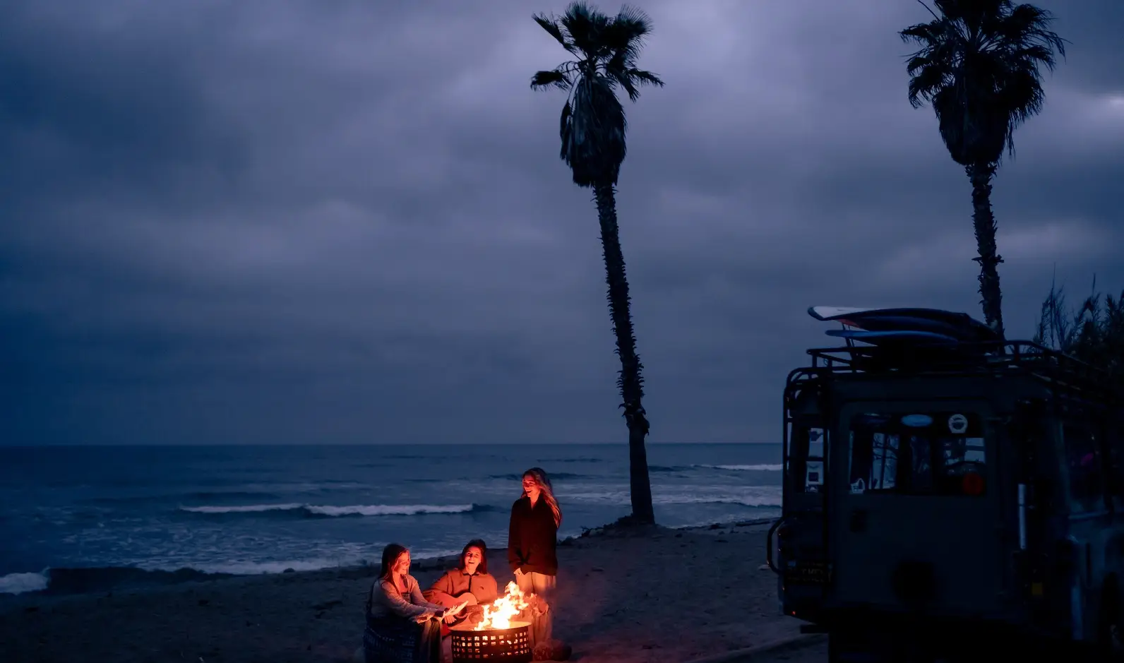 A soul community gathers around a campfire on a beach at dusk. The sky is overcast, and two tall palm trees are silhouetted against the horizon. A vehicle with surfboards on top is parked nearby, adding to the laid-back atmosphere.