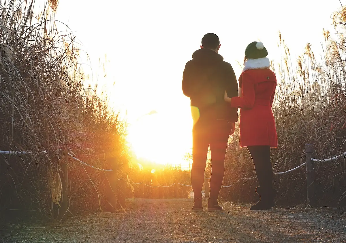 Two people, dressed warmly, stand side by side on a path flanked by tall grasses in Marin, looking towards the sunset. One wears a black jacket and the other a red coat with white fur trim. The golden sunlight bathes the scene, creating a warm, serene atmosphere perfect for discussing health tips.