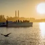A group of people enjoy outdoor activities in Monterey, standing on a boat cruising a tranquil lake during sunrise or sunset. The sun shines brightly in the sky, casting reflections on the water. A bird flies close to the water's surface, and two tall industrial chimneys are visible in the background.