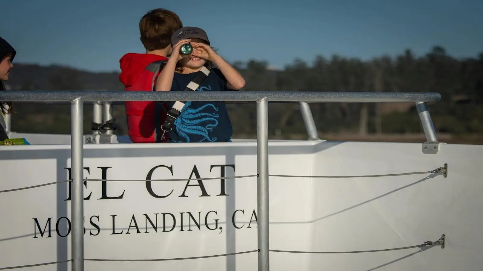 A child looks through binoculars while standing on a boat. An adult in a red jacket stands behind the child. Part of the boat's name and location, "EL CAT" and "MOSS LANDING, CA," are visible on the side. Trees and a clear sky are in the background, showcasing perfect family activities in Monterey Peninsula.