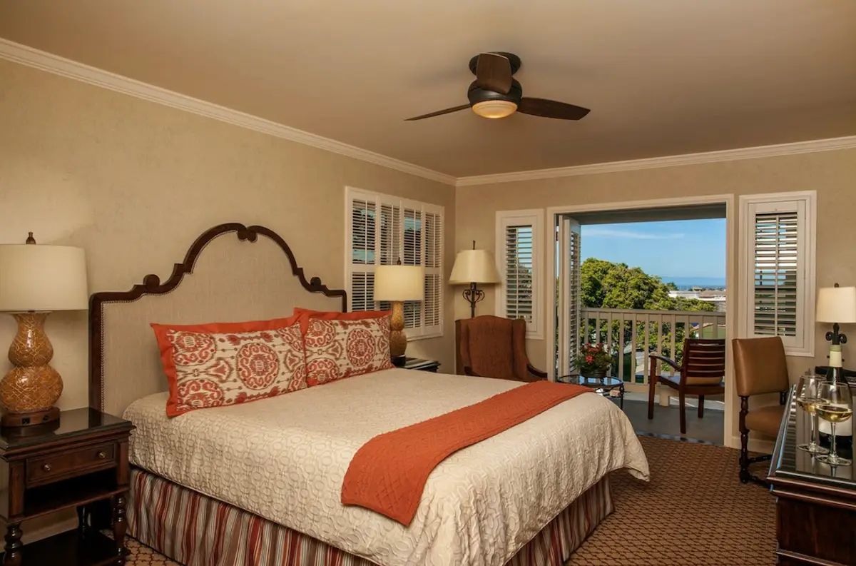 A cozy hotel room with a large bed decorated with pillows and an orange throw. The room features a ceiling fan, two armchairs, a small round table, and a desk. French doors open to a balcony with outdoor furniture offering a scenic view of lush greenery and the ocean—perfect for group travel in one of the best hotels in Monterey.