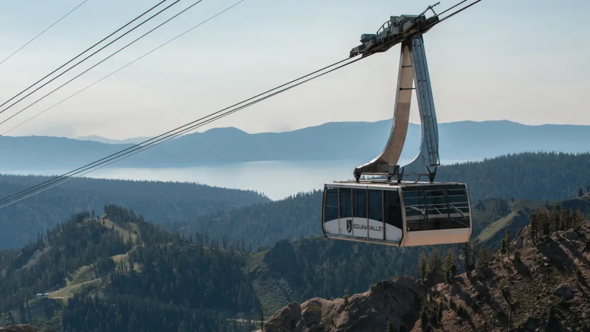 A cable car with large windows moves along a cable high above a mountainous landscape. Below, a dense forest covers the rugged terrain, leading to a distant body of water partially obscured by mist or haze—a perfect scene from a Tahoe summer guide.