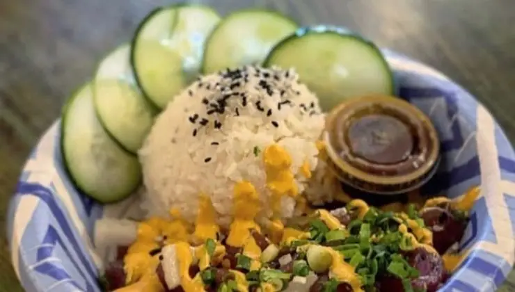 A bowl of food from Best Poke Central Kauai features sliced cucumbers, a mound of white rice topped with black sesame seeds, and a serving of diced fish or meat covered in creamy yellow sauce and chopped green onions. A small container of brown sauce is placed on the side.