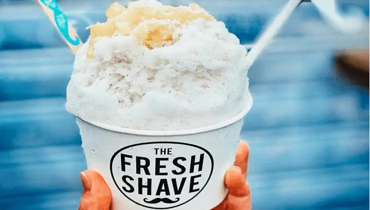 A hand holds a cup of shaved ice with a label that reads "The Fresh Shave." This best shave ice from Poipu is topped with pineapple chunks, and a spoon is placed in the cup. The background features blurred blue steps, reminiscent of Koloa and Waimea's charm.