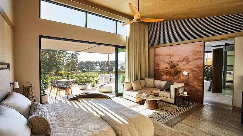 A modern, sunlit bedroom with large windows, a comfy bed, and a cozy seating area with a couch and coffee table. The room opens to an outdoor patio with table and chairs overlooking a green garden. A ceiling fan hangs from the wooden ceiling, adding rustic charm reminiscent of Napa hotels.