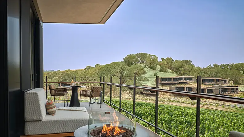 A balcony with a cozy seating area and fire pit in one of the finest Napa hotels overlooks a picturesque vineyard. The scene features lush green grapevines, modern buildings in the distance, and rolling hills under a clear blue sky. A table with chairs is also on the balcony.