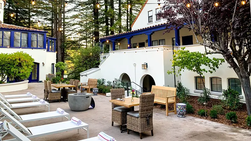 A charming outdoor patio with wooden tables and cushioned chairs is surrounded by lush greenery and string lights. White buildings with blue trim stand in the background, reminiscent of Napa hotels, and lounge chairs are arranged poolside under a canopy of tall trees.