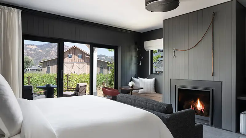 A modern bedroom akin to those in Napa hotels features a cozy fireplace, a large bed with white linens, and a sofa. There is a small dining table with two chairs near floor-to-ceiling windows that provide a view of a rustic barn and greenery outside. The room has dark walls and sleek decor.