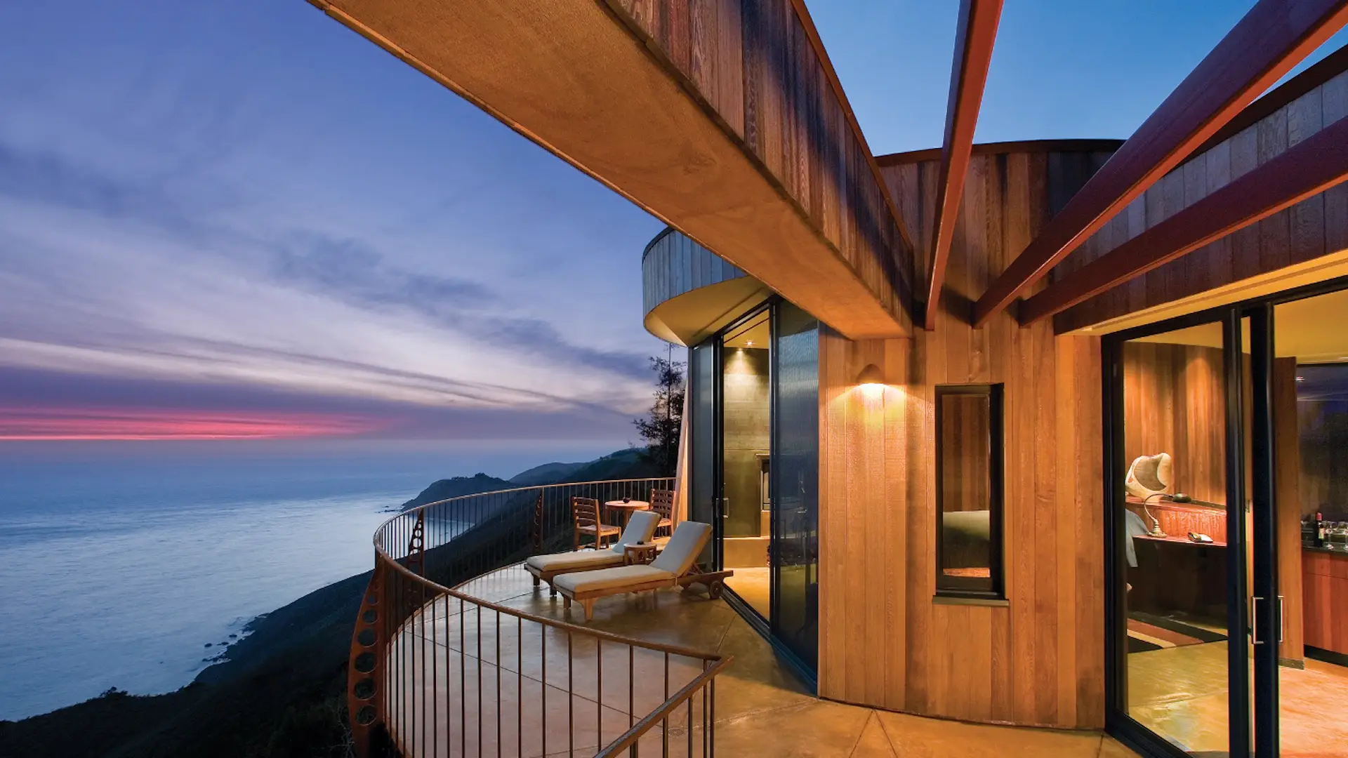 A modern wooden house with large windows and a curved balcony overlooks an expansive ocean landscape at sunset. Reminiscent of Big Sur romantic hotels, two lounge chairs are placed on the balcony, providing a relaxing spot to enjoy the serene view. The sky is painted with hues of purple and orange.