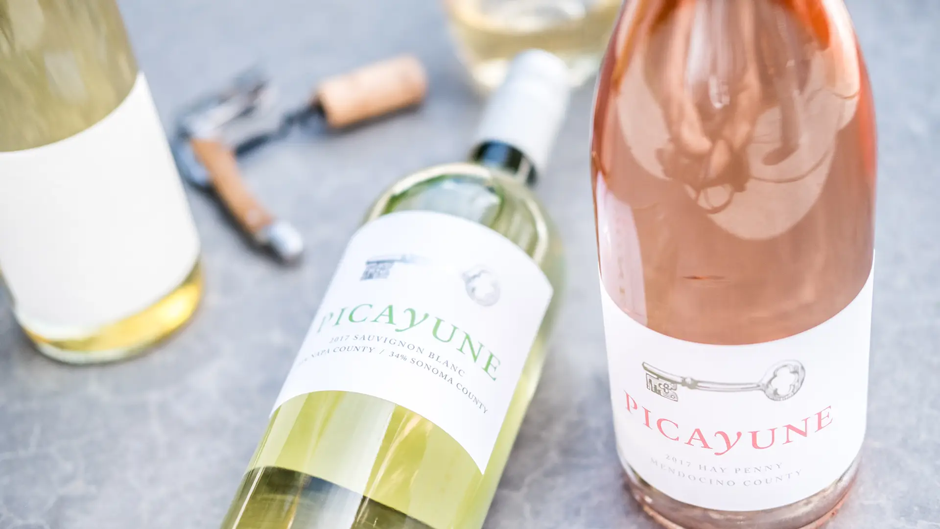 Two wine bottles labeled "Picayune" sit on a gray surface. One bottle, a 2017 Sauvignon Blanc from Sonoma County, has a green label, while the other, a 2017 Rosé from Mendocino County, dons a pink label. A corkscrew and cork lie nearby, evoking the charm of Napa Valley Tasting Rooms.