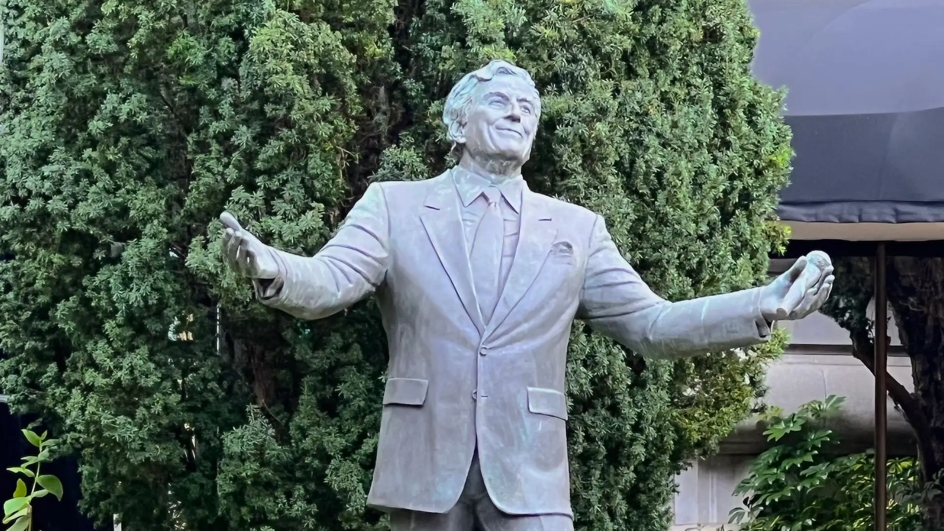 A bronze statue of a smiling man in a suit and tie with arms outstretched, resembling Tony Bennett, stands proudly. Set against a backdrop of dense, green foliage reminiscent of San Francisco's parks, the man's expression is welcoming and joyful.