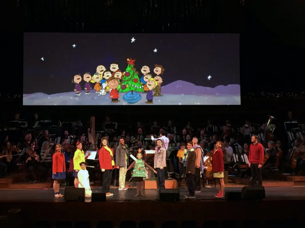 A group of performers on stage, dressed as characters from the Peanuts series. Behind them, a large screen displays an image of Peanuts characters around a Christmas tree, making it one of the delightful Bay Area holiday activities. An orchestra is seated in the background, indicating a musical performance.