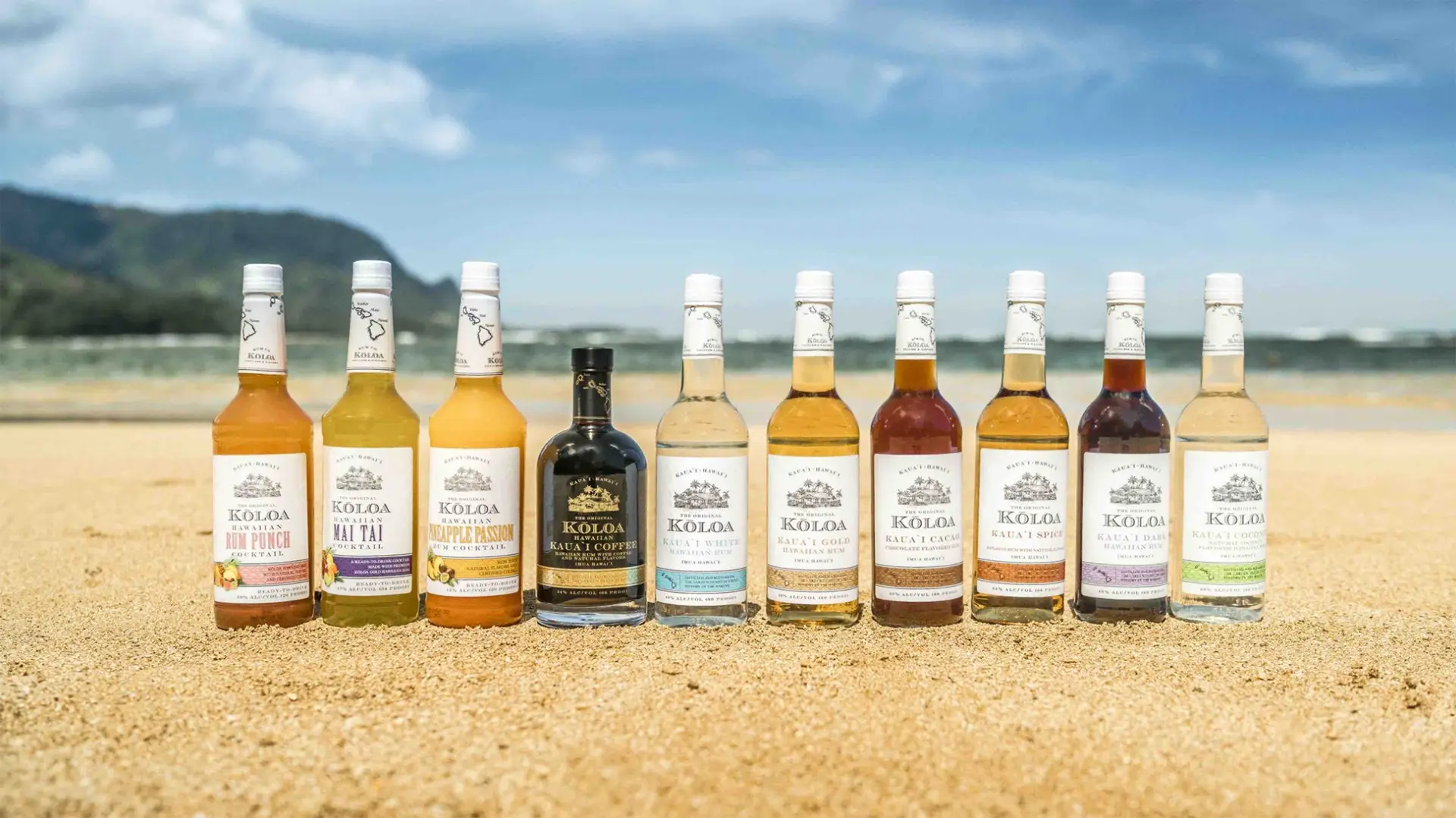 A lineup of 11 Koloa Rum bottles is displayed on a sandy beach, one of the best places to visit on Kauai. The bottles feature different colored labels, indicating various flavors. Blue sky and distant mountains enhance the idyllic background.