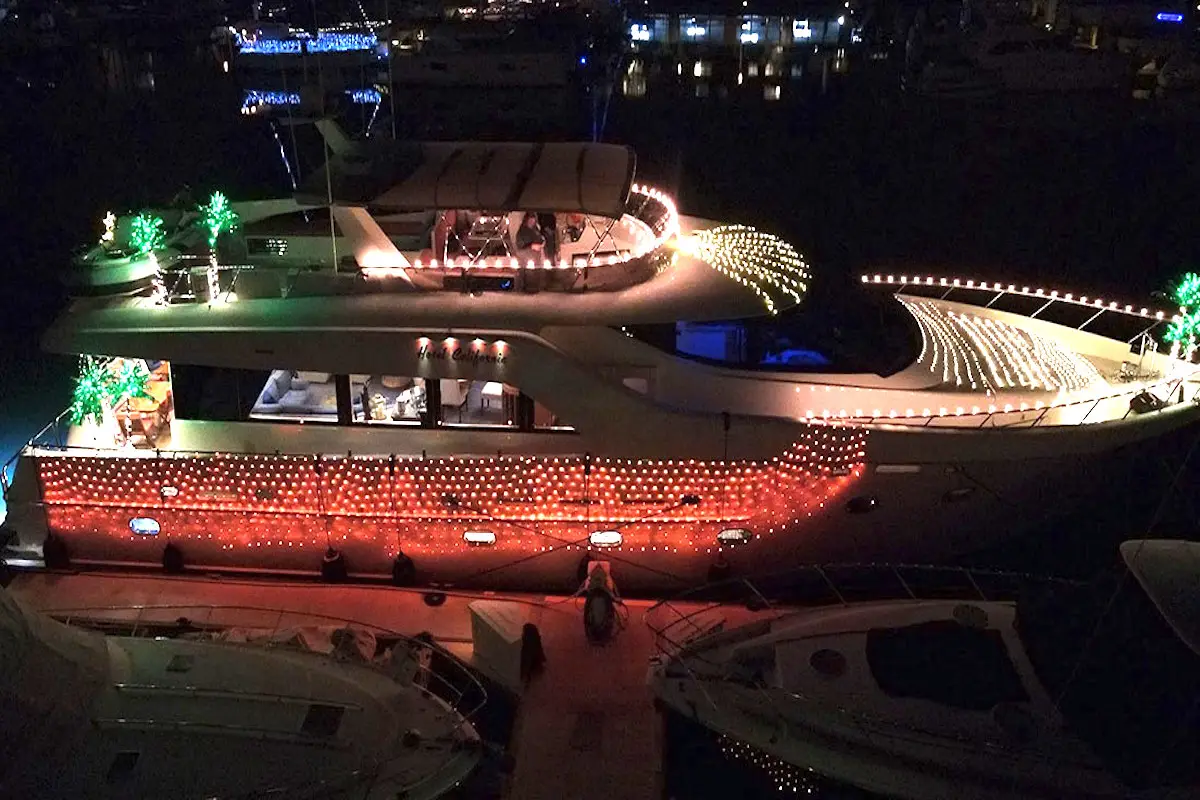 A large yacht is docked at a marina at night, illuminated with festive lights. The boat is adorned with strings of red, green, and white lights, and has additional lights outlining its shape. There is a small bridge connecting it to the dock. This dazzling display is one of the Bay Area holiday activities.