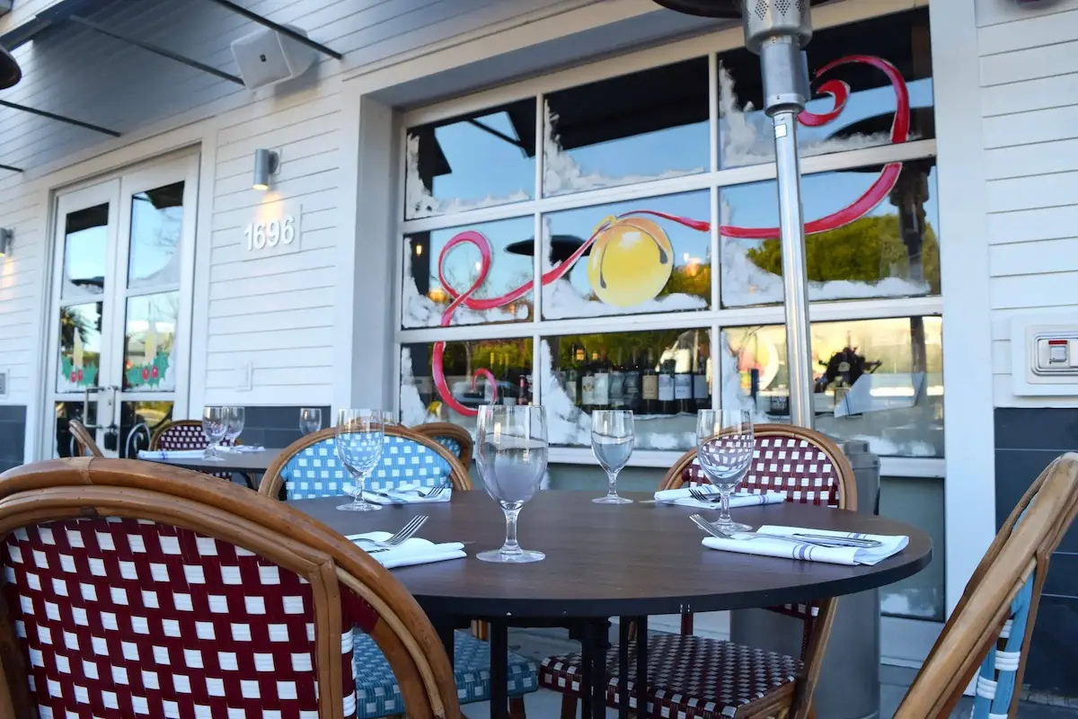 Outdoor seating area at a café with a round table set with empty wine glasses and folded white napkins. The chairs have woven red and blue patterns. Behind the table is a window decorated with a bright yellow sun and red swirls, reflecting outdoor trees—perfect for enjoying Bay Area holiday activities.