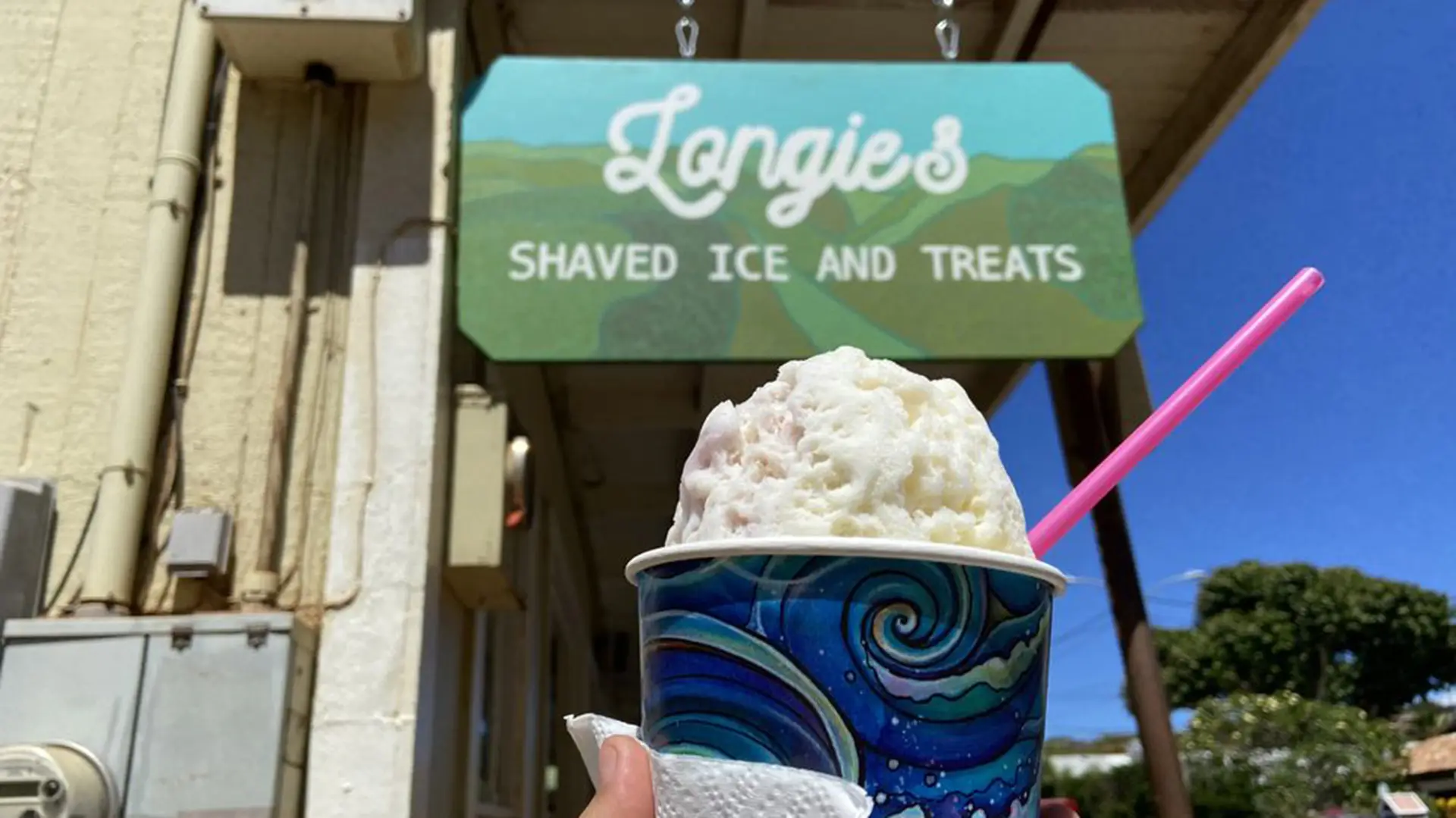 A hand holding a cup of the best shaved ice in Poipu, with a blue and green swirled pattern backdrop, a napkin, and a pink spoon. In the background, a sign reads "Longie's Shaved Ice and Treats" featuring a mountain design. It's a bright, sunny day with clear skies.