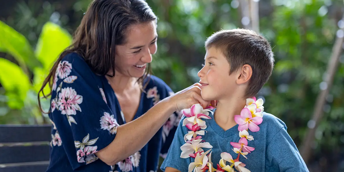 A woman in a floral blouse smiles as she places a lei around the neck of a smiling boy in a blue shirt. They are outdoors, surrounded by lush greenery.