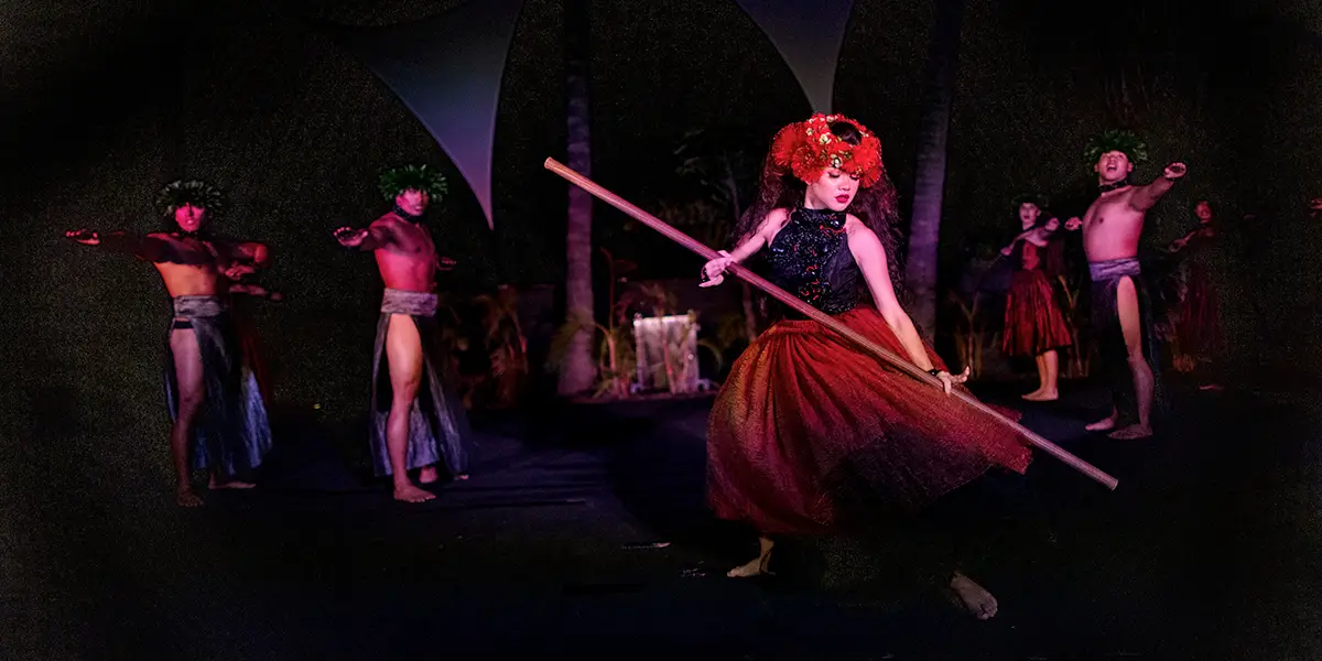 A group of people perform a traditional Polynesian dance on stage. In the foreground, a person dressed in a red skirt and floral headpiece holds and balances a large wooden staff. Other dancers in red and traditional attire are seen in the background.