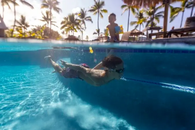 Underwater view of a boy swimming in a pool wearing goggles, with another child standing near the edge of the pool. Bright sunlight and palm trees are visible in the background, creating a tropical atmosphere perfect for Kona teenager activities.