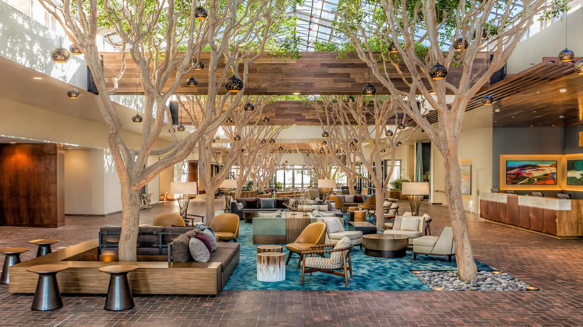 A luxurious hotel lobby in Monterey boasts a high glass ceiling and large indoor trees, creating a natural ambiance. The space is furnished with modern seating arrangements in various styles and colors, centered on a blue rug. Warm lighting enhances the inviting atmosphere, offering value beyond typical hotels.