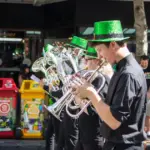 A band of musicians wearing bright green hats and black outfits performs outdoors, celebrating St. Patrick’s Day. They play a variety of brass instruments. In the background, several waste bins in different colors and a group of onlookers are present. Sunlight illuminates the festive scene.