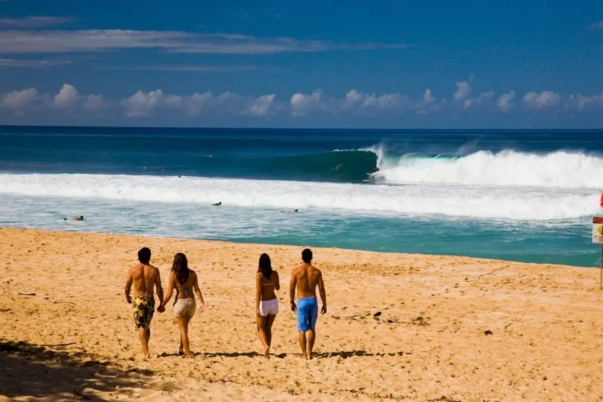 Four individuals, two men and two women, are walking on a sandy beach towards the ocean. The sky is clear with some clouds on the horizon. The waves are moderate, and a few surfers can be seen in the water. The group appears relaxed and enjoying one of the best beaches for long walks on Oahu.