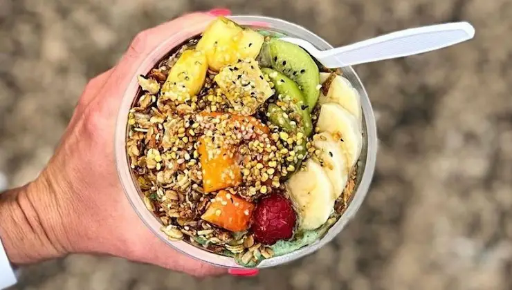 A hand holding a bowl filled with a colorful assortment of fruit including banana slices, kiwi, pineapple, raspberries, and papaya. The bowl also contains granola and seeds, with a white plastic spoon on the side—reminiscent of the vibrant smoothies and acai bowls from Kauai.