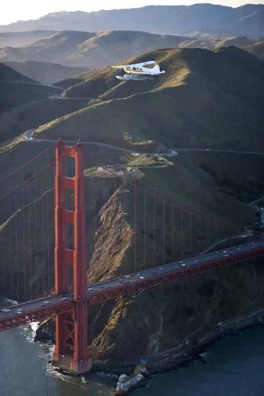 An aerial view of the Golden Gate Bridge with a seaplane from Seaplane Adventures flying above it. The iconic red-orange suspension bridge spans across the water, with rolling hills in the background under a clear sky. Cars are visible traveling along the bridge.