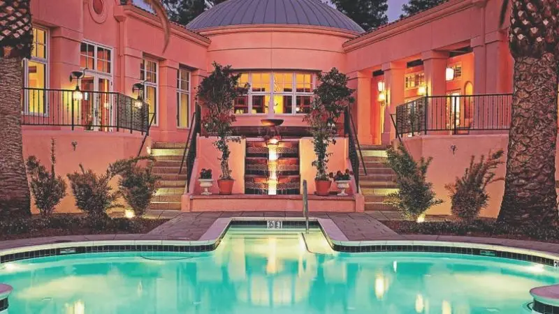 A luxurious mansion with a pinkish exterior features a grand staircase leading to a central domed structure. Potted plants line the stairs, and a small fountain cascades down the steps. At the forefront is a turquoise, illuminated swimming pool, surrounded by greenery.