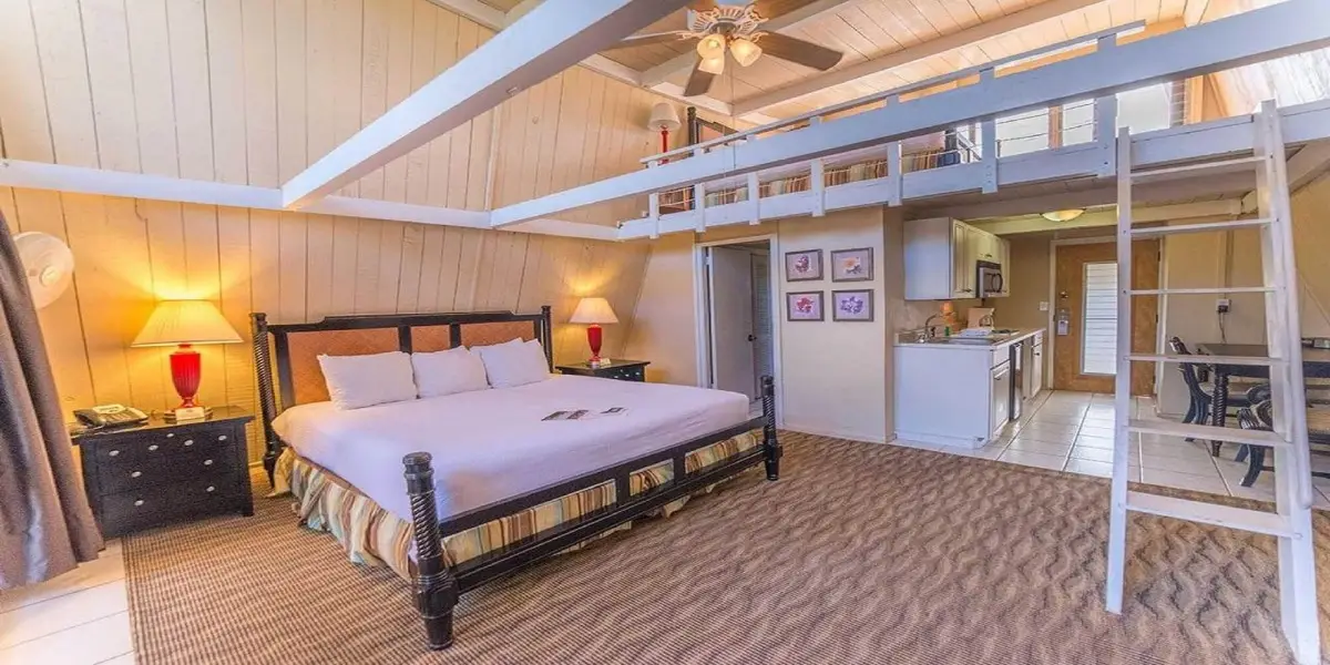 A cozy, loft-style bedroom with a large bed, two bedside lamps, and a striped rug. A wooden ladder leads to an upper loft area. An open kitchen with white cabinets, a dining table and chairs are visible in the background. Walls are light wood paneling—ideal for those seeking value hotels Maui.