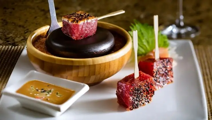 A gourmet dish featuring seared tuna cubes on wooden skewers, seasoned and partially cooked. A black hot stone for further cooking sits in a wooden bowl. A small white dish contains a dipping sauce, all presented on a white rectangular plate. Experience the best gluten-free dining in Wailea.